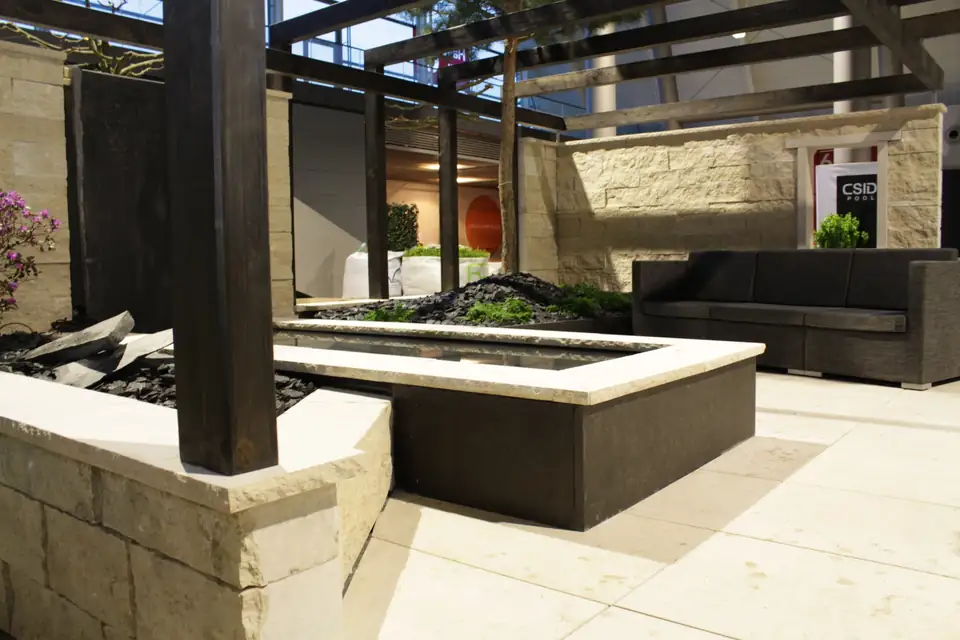 Wasserbecken mit Natursteinrand unter der Laube, dahinter Lounge-Sofa und Natursteinwand